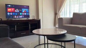 Un salon moderne avec un téléviseur à écran plat fixé sur un meuble de télévision en bois foncé. On y trouve une table basse ronde en verre au centre, un canapé gris à gauche et un canapé beige à droite. La pièce est dotée d’une grande fenêtre avec des rideaux légers.
