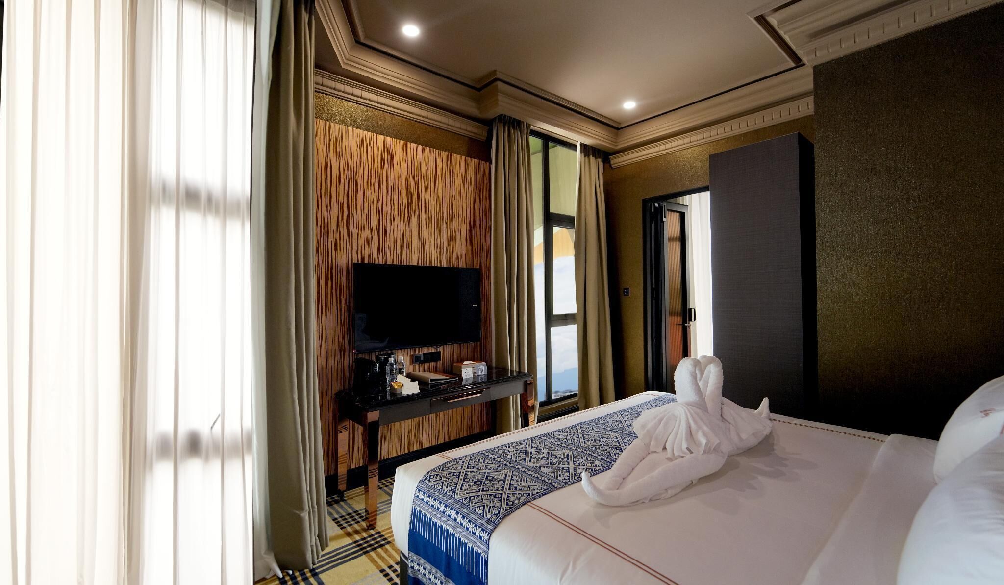 A hotel room with a large bed, a flat-screen TV mounted on a wooden panel wall, a bedside table with a coffee maker, and a view of the outdoors through a window.