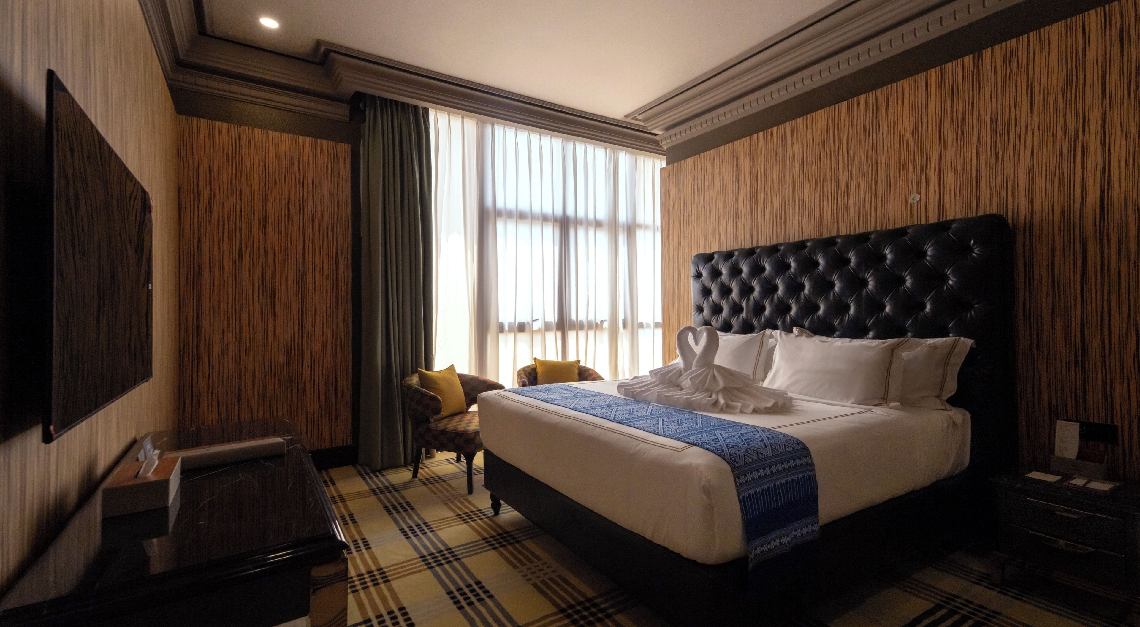 Een hotelkamer met een groot bed, een flatscreen televisie, een wand van houten panelen en een raam met gordijnen.