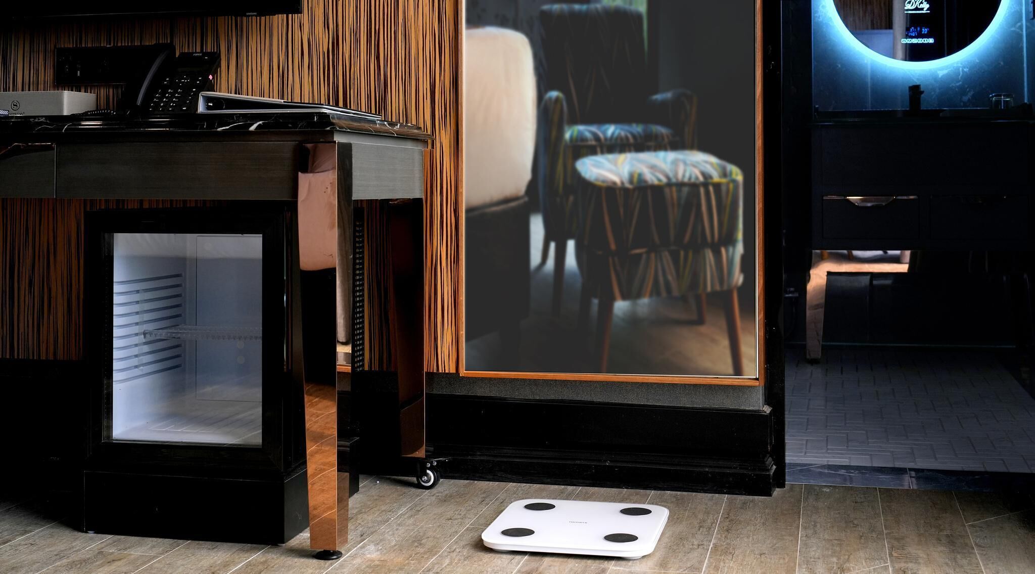 A modern room with a large mirror reflecting a chair and a sofa, a desk with a phone, and a scale on the floor.