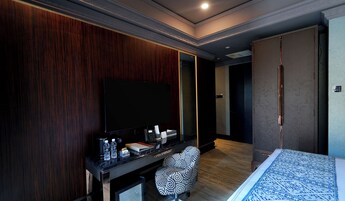 Ett modernt hotellrum med en stor platt-TV, en väggpanel i trä, ett skrivbord med en kaffebryggare, vattenflaskor och en stol med ett mönstrat tyg.