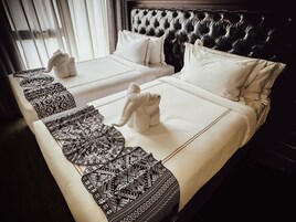 A hotel room with two beds, each with a white bedspread and matching pillows. There are two towels placed on each bed. The room has a dark headboard and a window with curtains.