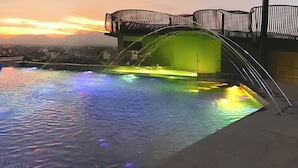 An infinity pool with a view of a cityscape at sunset, a glass railing, and a water feature. - 1969 DKelly Hotel  (Ipoh)