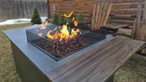 Ein rechteckiges Feuerbecken im Freien mit Glasplatte und einem Holztisch davor.