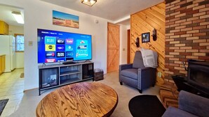 Ein Wohnzimmer mit Couchtisch aus Holz, einem Fernseher, der verschiedene Sender zeigt, und einem Kamin.