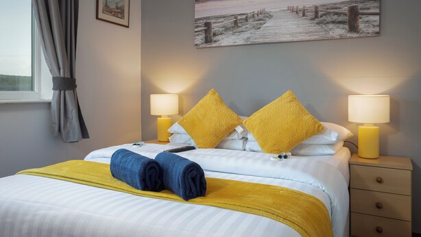A neatly made bed with yellow and blue towels, two bedside lamps, a framed picture on the wall, and a window with curtains. - Royal Oakwell Hotel (Blackpool)