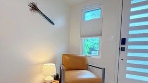 Une chambre moderne avec un fauteuil en cuir, une petite table en bois surmontée d’une lampe et une fenêtre équipée de stores.
