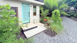 Une maison moderne avec une porte turquoise, un petit porche et un jardin où poussent différentes espèces de plantes.