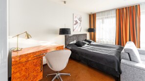 Et hotelværelse med et træskrivebord, en moderne stol, en seng med gråt sengegavl og et vindue med orange gardiner.