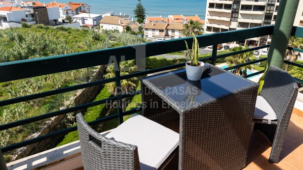 Een balkon met rieten meubilair, een plant in pot en uitzicht op zee en gebouwen.
