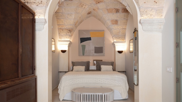 Une chambre avec une arche en pierre, un lit recouvert d’une couette blanche, une table ronde et deux lampes fixées au mur.