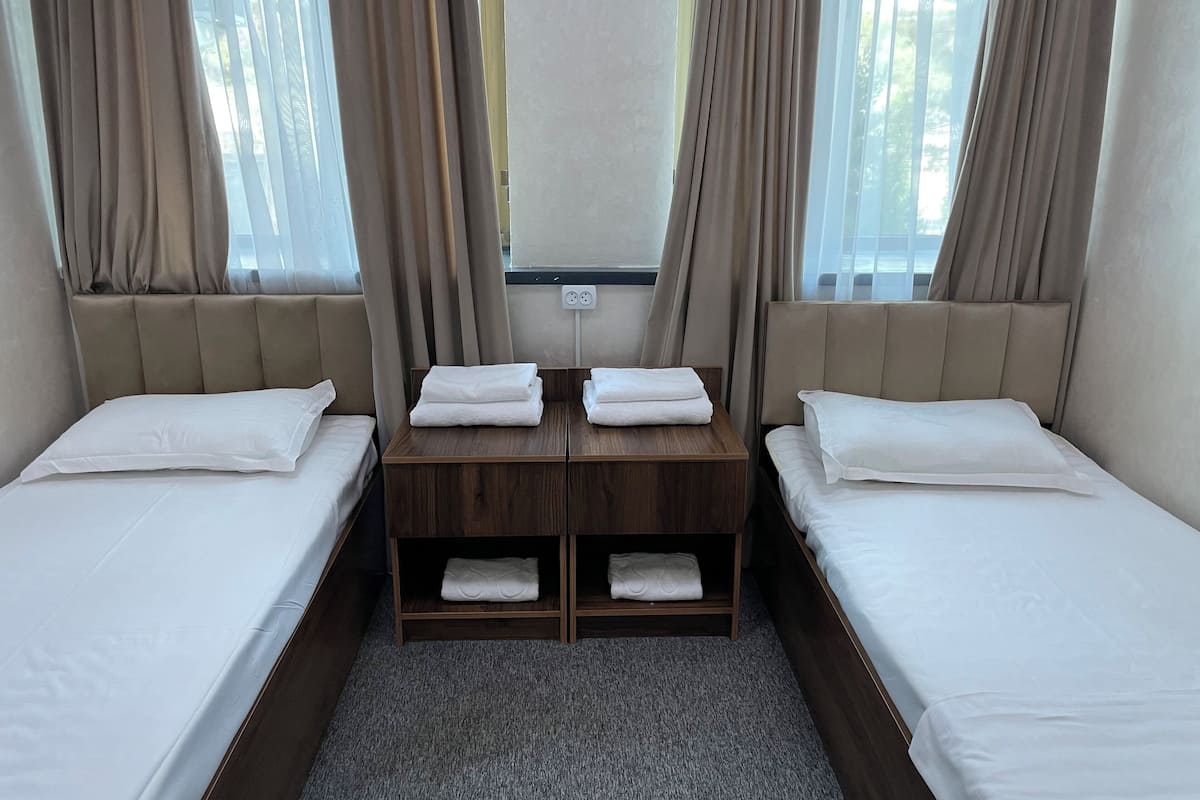 Une chambre d’hôtel avec deux lits simples, une table de chevet en bois avec des serviettes et une fenêtre avec des rideaux.