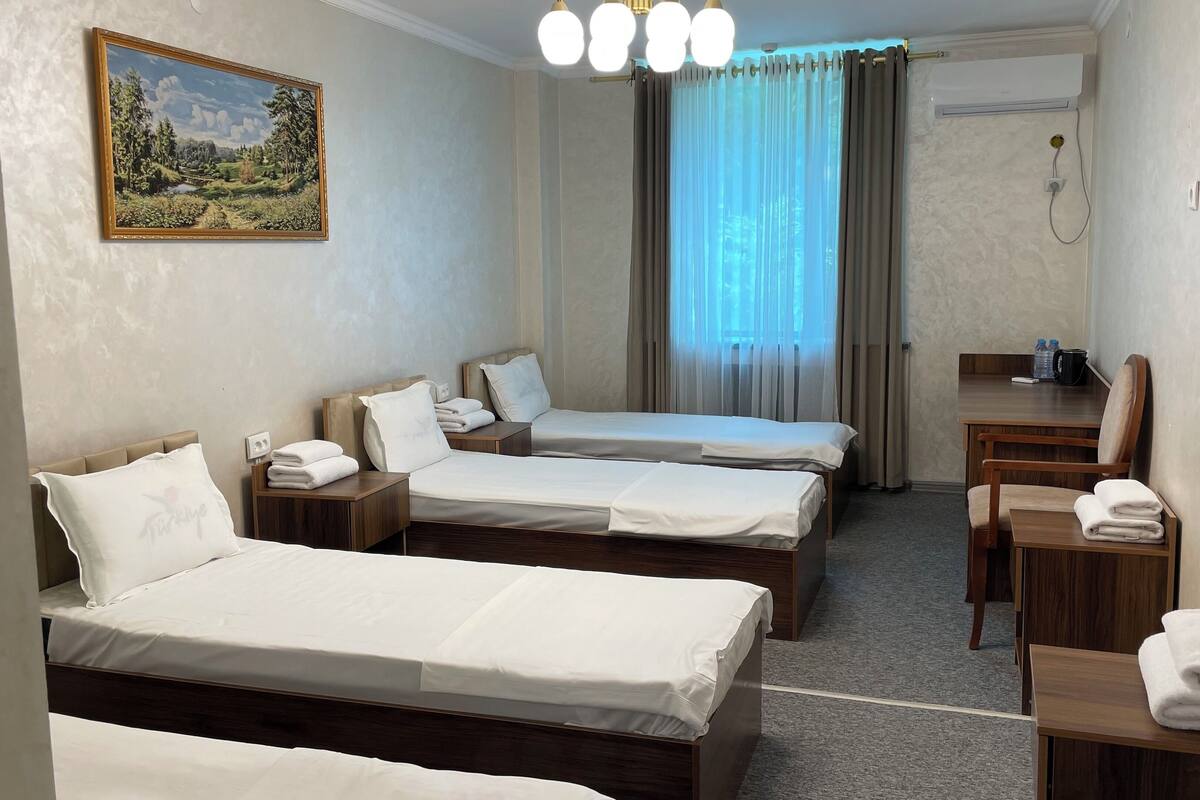 Une chambre d’hôtel avec deux lits simples, un bureau avec une chaise, une petite table avec une chaise, un tableau au mur et une fenêtre avec des rideaux.