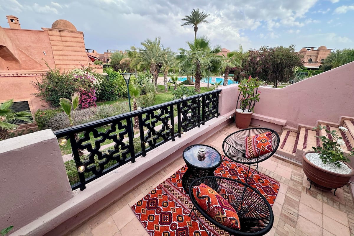 A terrace with a patterned rug, two round tables with cushions, potted plants, and a view of a swimming pool and palm trees.