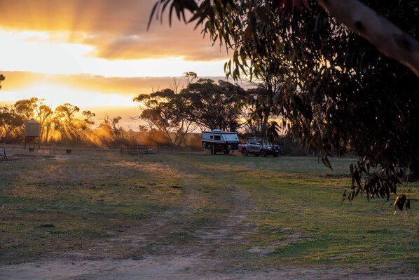 A rural landscape with a dirt path leading to a white RV parked on grass, surrounded by trees and a sunset in the background.