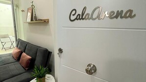 Un salón moderno con un sofá gris, una pequeña mesa blanca con una planta, y una puerta con la etiqueta 'CalaArena'.