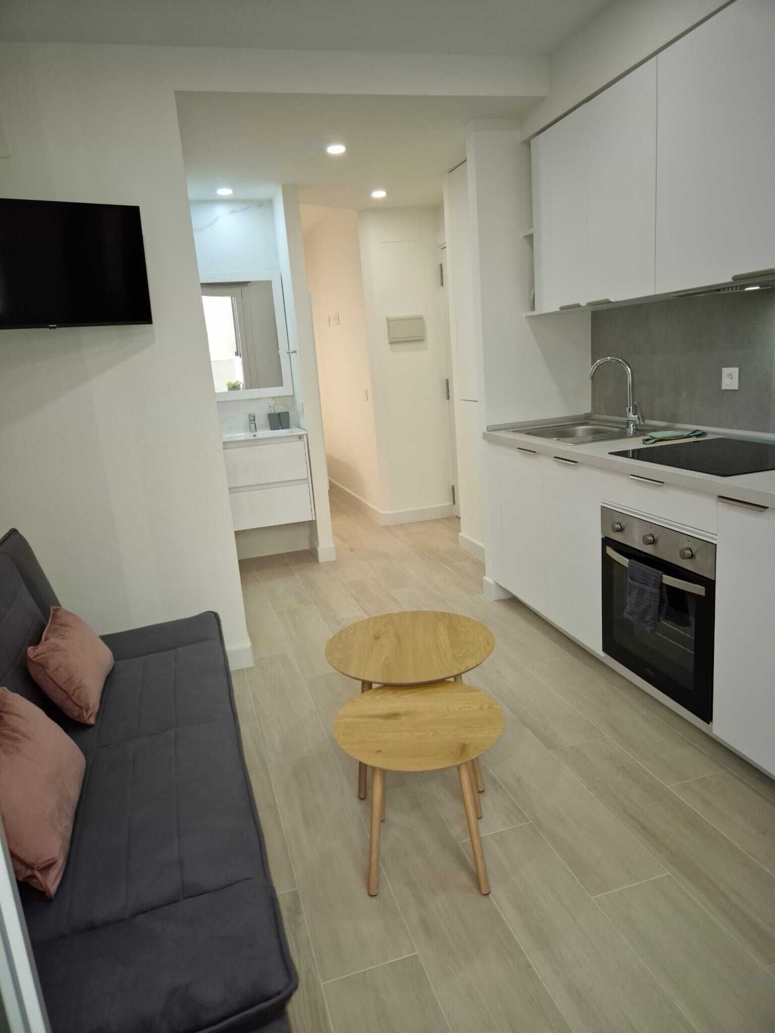 Cocina compacta con armarios blancos, horno empotrado y pileta. Hay una pequeña mesa de madera y un sofá oscuro con almohadones.