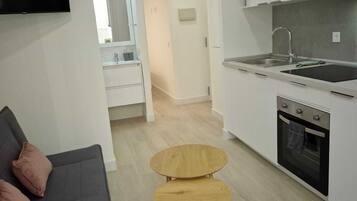 Cocina compacta con armarios blancos, horno empotrado y pileta. Hay una pequeña mesa de madera y un sofá oscuro con almohadones.