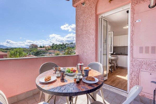 A balcony with a table set for breakfast, a view of the city, and a sliding door leading to a kitchen. - Villa Paolina Apartments (Ceriale)