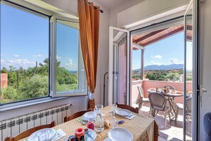 A dining area with a table set for two, a view of greenery and mountains through large windows, and an open door leading to an outdoor patio. - Villa Paolina Apartments (Ceriale)
