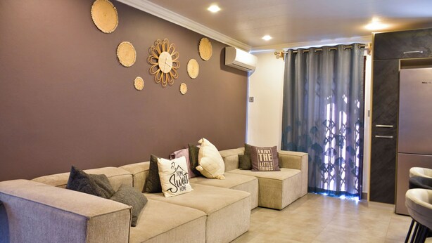 A modern living room with a beige sectional sofa, decorative wall art, and a kitchen area with a refrigerator.