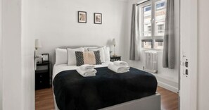 Una habitación de hotel con cama, mesita de noche, lámpara, ventana con cortinas y un radiador.