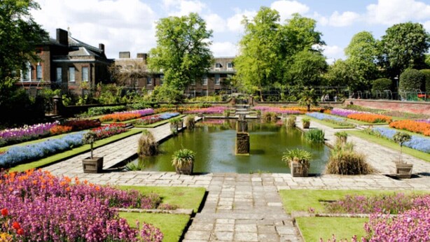 Un jardin à la française avec un bassin central, des parterres de fleurs soigneusement entretenus et un sentier en pierre menant à un bâtiment à la façade en briques.