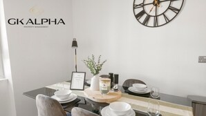 Un espace de salle à manger moderne avec une table dressée pour quatre personnes, agrémentée d’assiettes et de bols blancs, ainsi que d’un centre de table. On peut également voir une horloge murale et un certificat encadré.