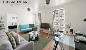 Una sala de estar moderna con un sofá gris, un sillón azul, una mesa de centro y un comedor con mesa dispuesta para dos.