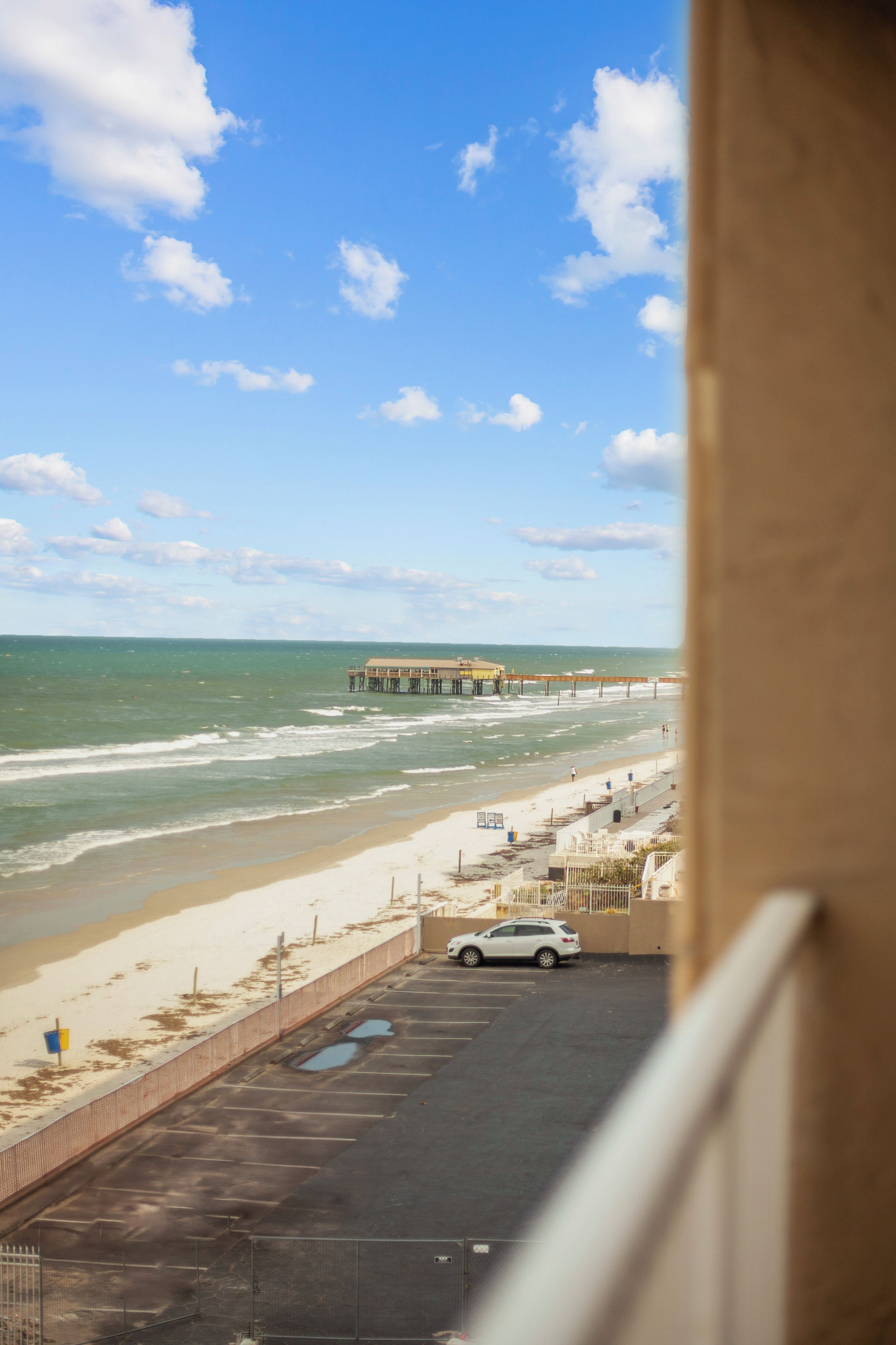 Blick auf einen Strand mit einem Steg, einem Parkplatz und einem Gebäude im Vordergrund.