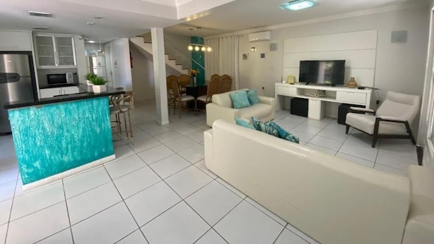 A modern living room with a white sofa, a kitchen area with a turquoise island, and a dining area with chairs and a table. - Parada Beach Apartamentos (Florianopolis)