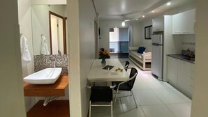 Een moderne keuken met witte kasten, een eethoek met een witte tafel en stoelen, en een zichtbare badkamer met een wit wastafelmeubel en spiegel.