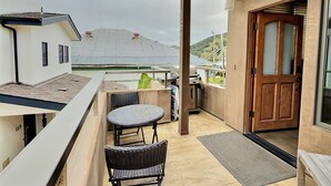 A balcony with a wicker table and chairs, a wooden door, and a view of a building with solar panels. - Sun 'n Sand Studio at Avila - a perfect beach spot! (Avila Beach)
