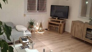 A living room with a TV, wooden furniture, a glass coffee table, and a potted plant. - Charming T3 villa near the sea (Le Pradet)