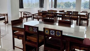 A conference room with a glass table surrounded by wooden chairs with colorful backrests, large windows with curtains, and a ceiling with recessed lighting. - Kala Retreat (Rishikesh)