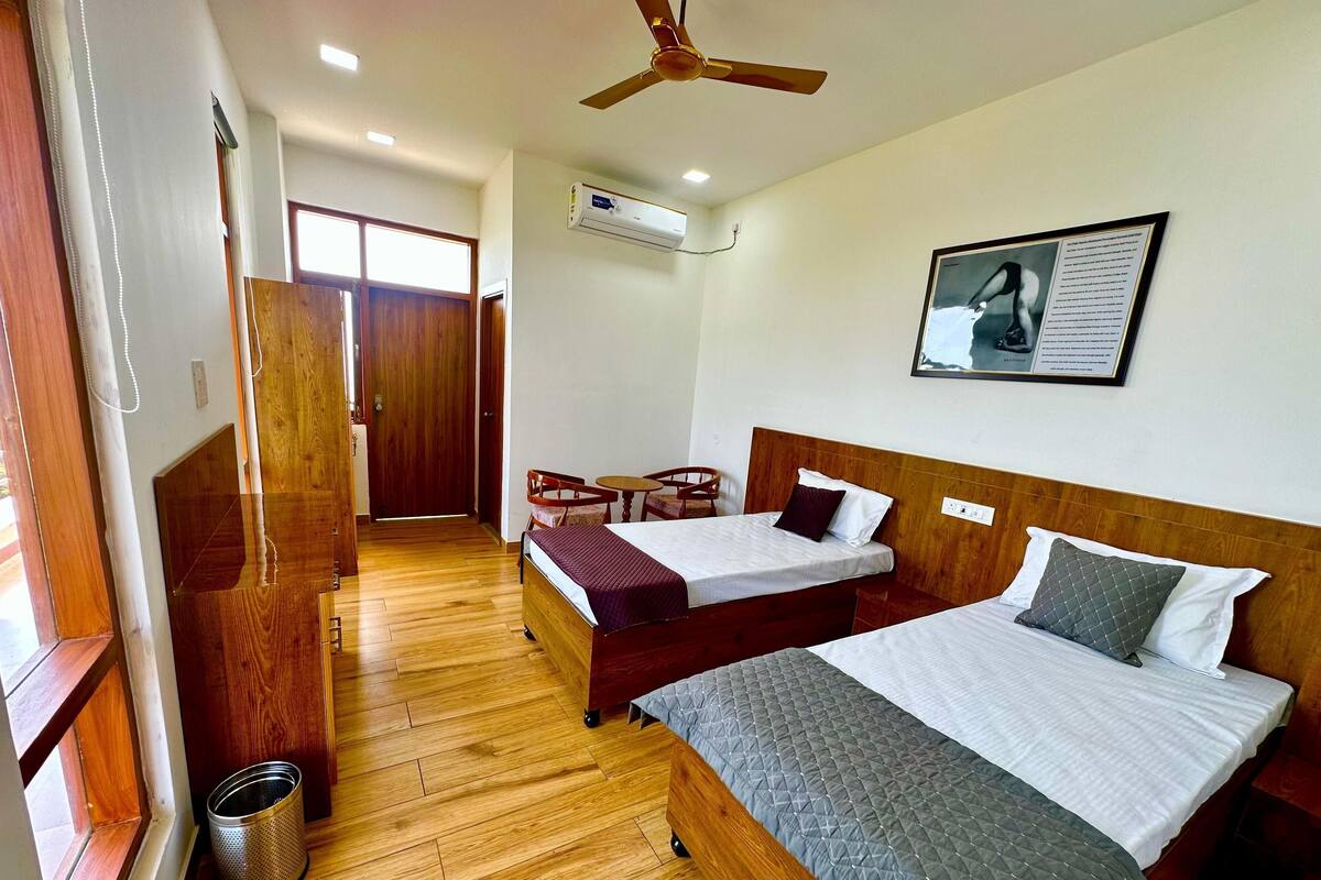Une chambre d’hôtel avec deux lits, un ventilateur de plafond, un appareil de climatisation, un tableau encadré au mur et un plancher en bois.