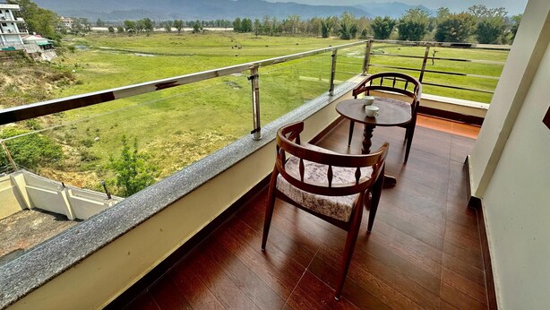 A balcony with a table and chairs, offering a view of a green field and distant mountains. - Kala Retreat (Rishikesh)