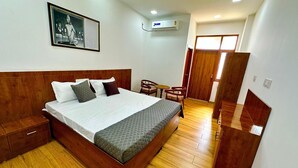 A neatly arranged bedroom with a wooden bed, a small table, and a chair, an air conditioning unit, and a door leading to another room. - Kala Retreat (Rishikesh)
