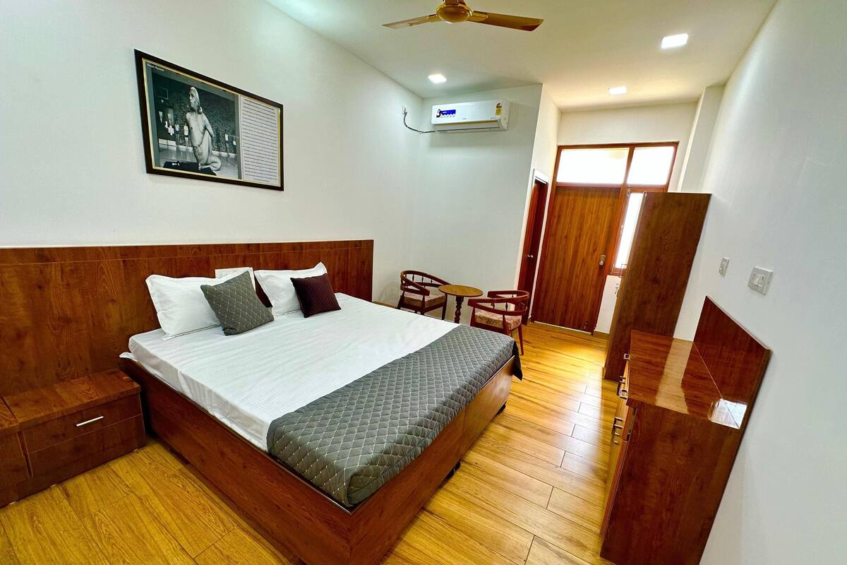 Une chambre à coucher bien rangée, avec un lit en bois, une petite table et une chaise, un appareil de climatisation et une porte donnant sur une autre pièce.