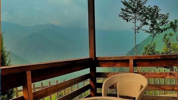 Une terrasse en bois, agrémentée de meubles en osier et d’une table en verre, qui offre une vue sur un paysage montagneux.