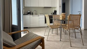 Una sala de estar moderna con sofá, mesa de comedor y sillas, una cocina con armarios blancos y un ventanal con cortinas.