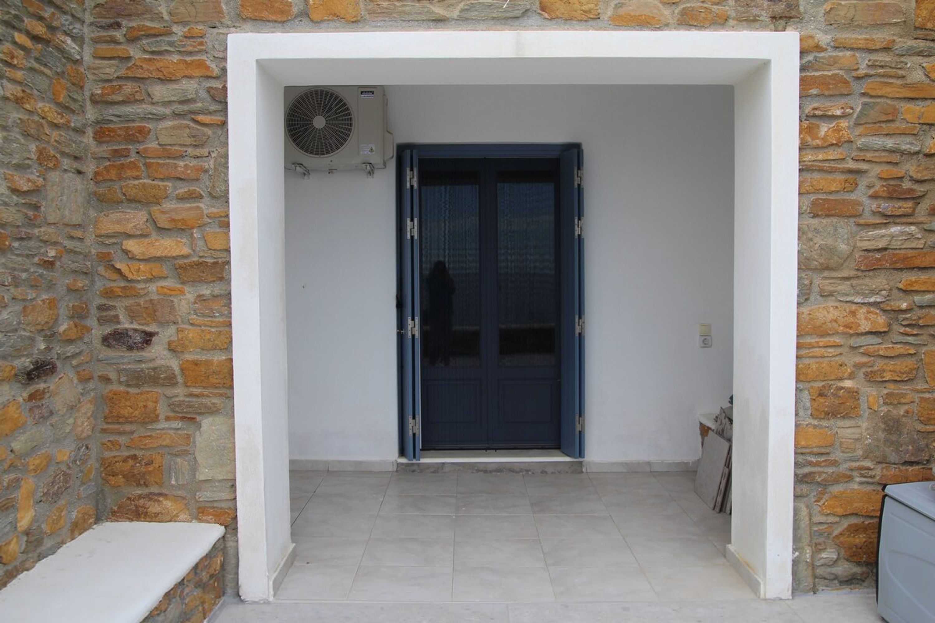 Una entrada de paredes de piedra con una puerta azul, un banco blanco y un ventilador fijado a la pared.