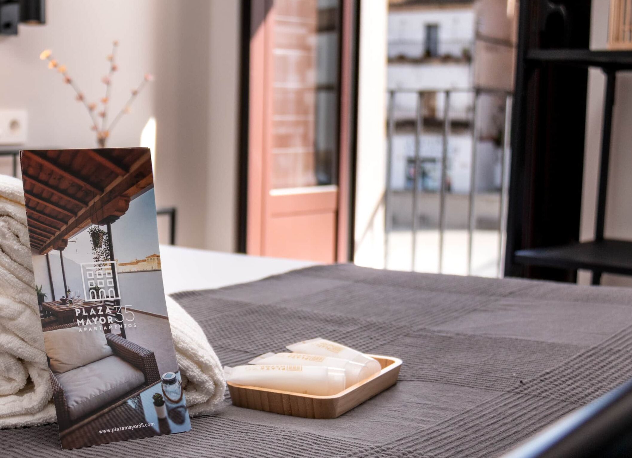 A neatly made bed with a brochure, a towel, and a wooden tray with a candle.