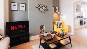 Una sala de estar moderna con un televisor de pantalla plana, una mesa de centro y un sofá. Se ve una cocina a través de un vano abierto.