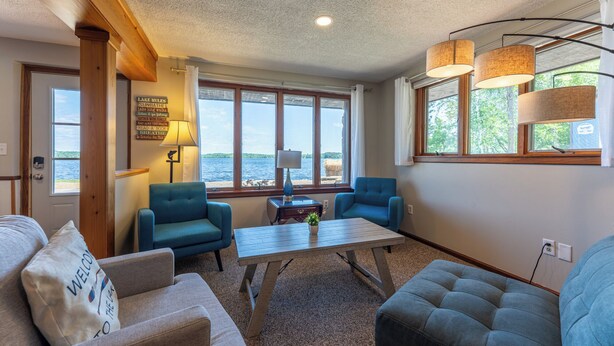 Una habitación acogedora con vistas a un lago, que cuenta con techo de madera, dos sillones azules, un sofá beige y una pequeña mesa con lámpara.