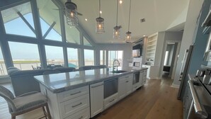Una cocina amplia con una isla grande, armarios blancos y vistas a la playa a través de amplios ventanales.