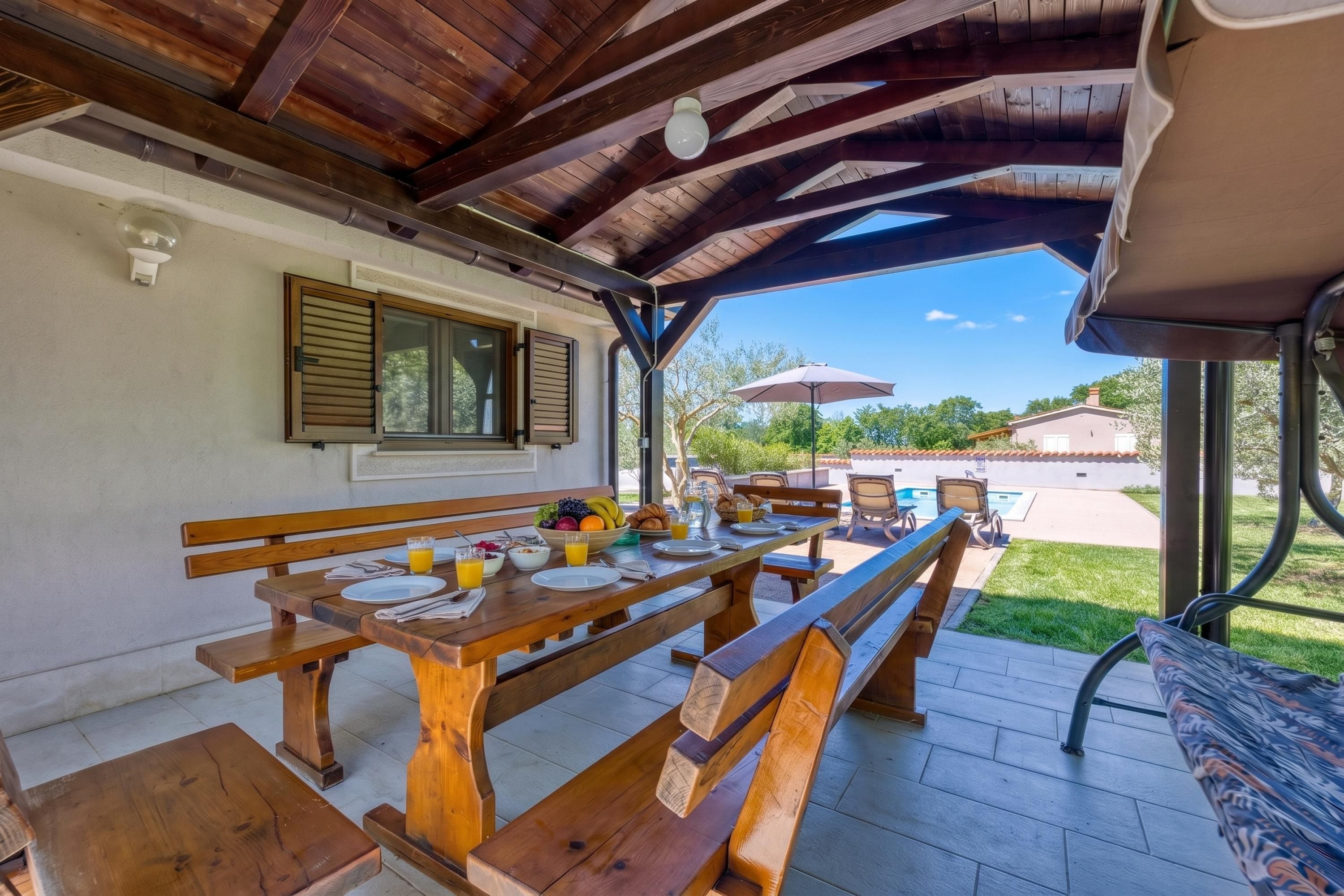 An outdoor dining area with wooden benches and tables, a pool in the background, and a clear sky.