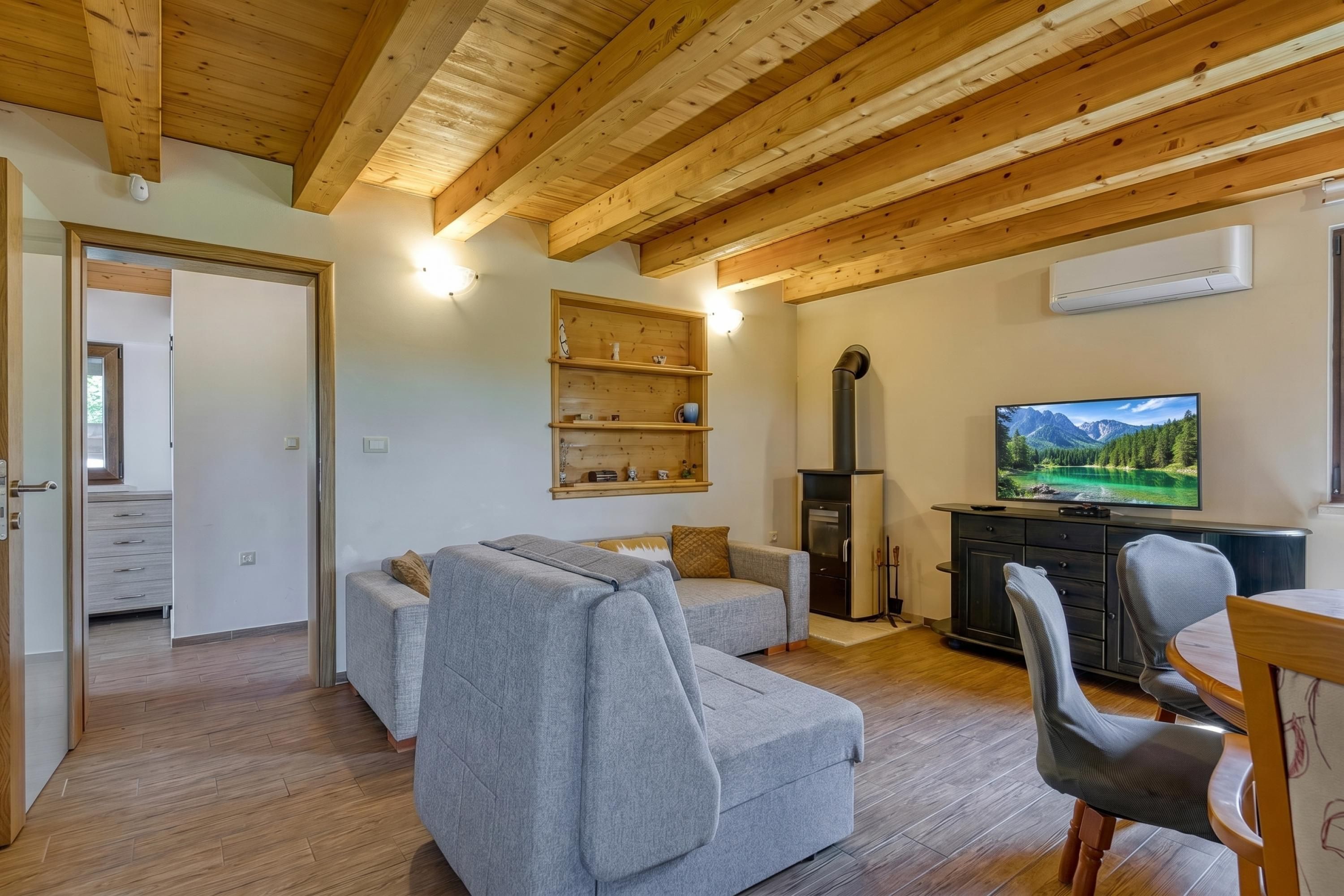 Een gezellige woonkamer met een grijze bank, houten balken in het plafond, een televisie aan de muur en een keuken in de achtergrond.