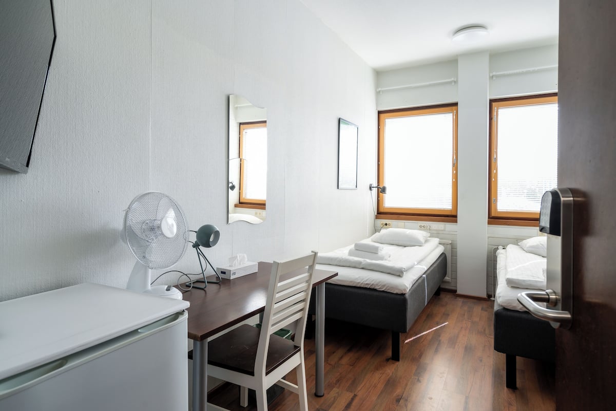 A compact hotel room with a desk, chair, two beds, a mirror, and a fan.