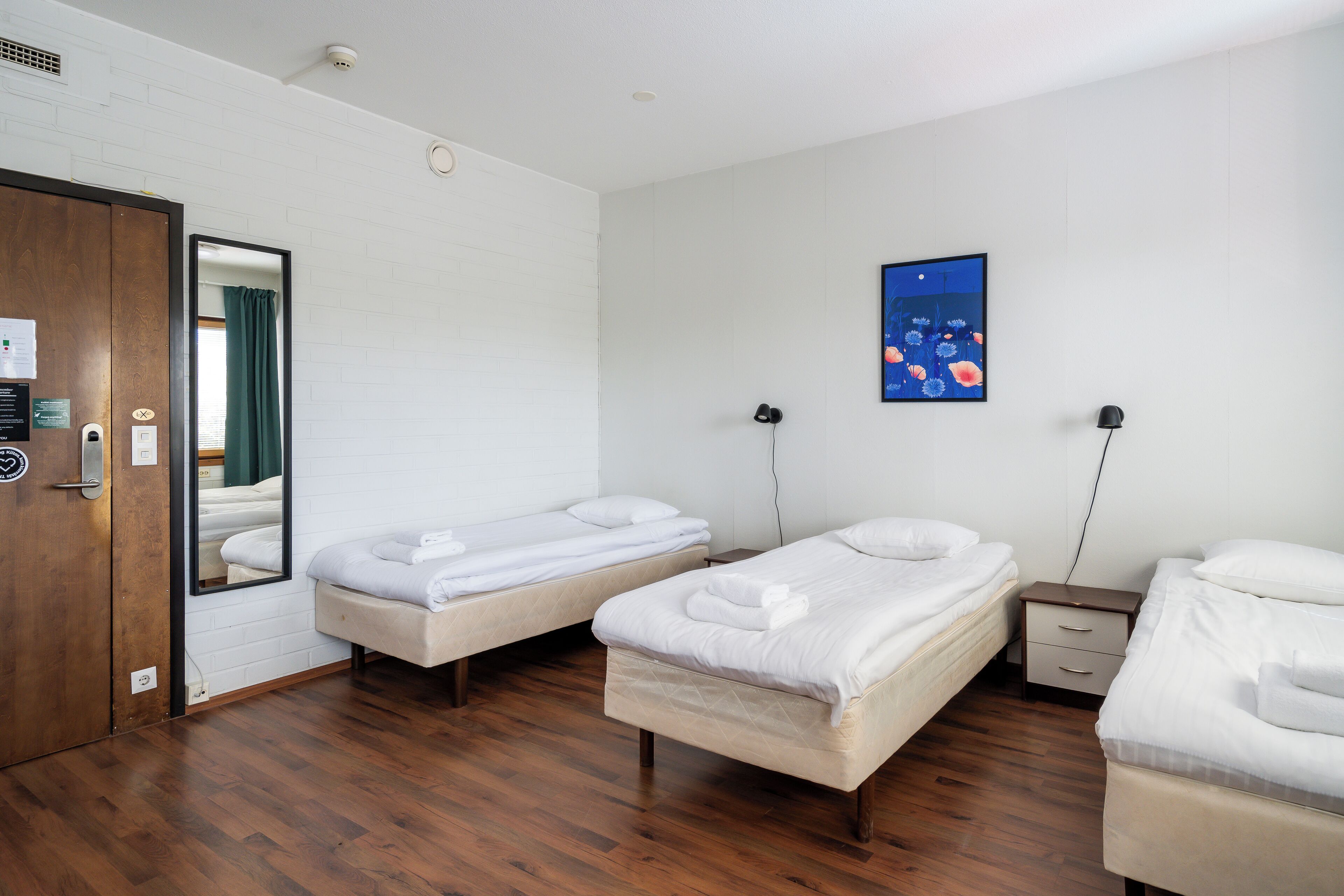 Habitació d'hotel amb dos llits individuals, un terra de fusta, un mirall, una tauleta de nit i una obra d'art penjada a la paret.